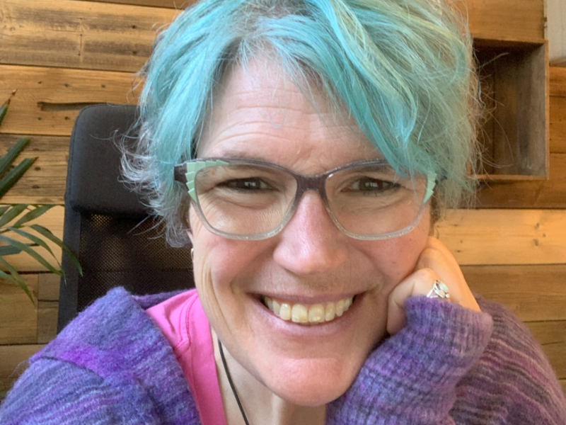 woman resting chin in hand with blue hair leaning on desk smiling